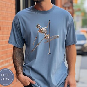 May include: Blue jean t-shirt featuring a skeleton figure pole dancing. The skeleton is depicted in a dynamic pose, holding onto a silver pole. The shirt has a round neck and short sleeves. The text "BLUE JEAN" is in a circle on the bottom left.