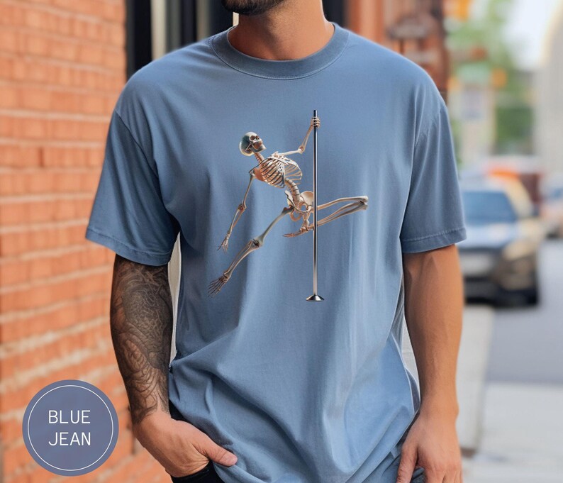 May include: Blue jean t-shirt featuring a skeleton figure pole dancing. The skeleton is depicted in a dynamic pose, holding onto a silver pole. The shirt has a round neck and short sleeves. The text "BLUE JEAN" is in a circle on the bottom left.