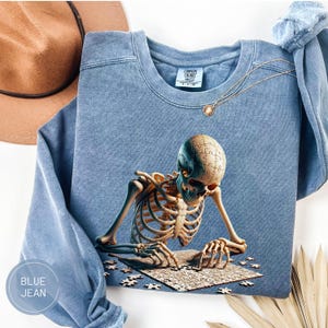 May include: Blue jean-colored sweatshirt featuring a detailed illustration of a skeleton assembling a puzzle. The puzzle pieces are scattered around the skeleton, and the sweatshirt has a crew neck and long sleeves. The text "BLUE JEAN" is in a circle on the bottom left.