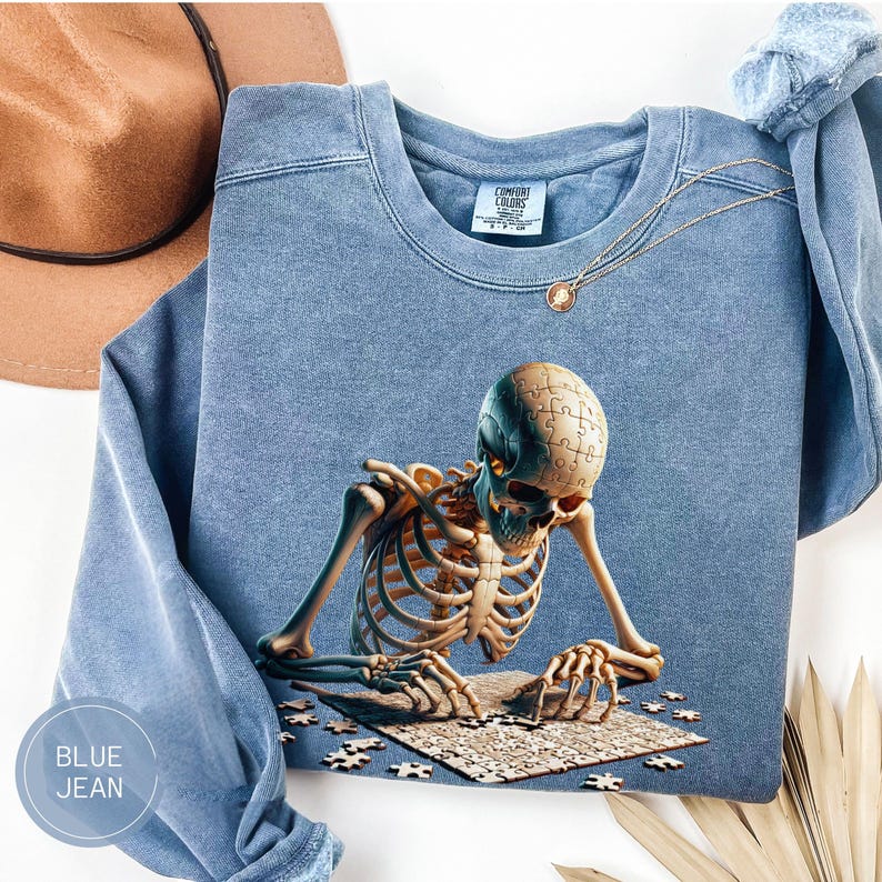 May include: Blue jean-colored sweatshirt featuring a detailed illustration of a skeleton assembling a puzzle. The puzzle pieces are scattered around the skeleton, and the sweatshirt has a crew neck and long sleeves. The text "BLUE JEAN" is in a circle on the bottom left.