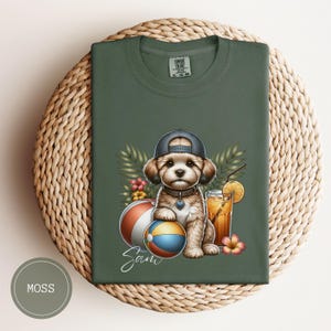 May include: A green t-shirt with a cartoon illustration of a dog wearing a blue baseball cap and holding a beach ball. The dog is surrounded by tropical plants and a glass of orange juice. The text "Sam" is written below the dog.