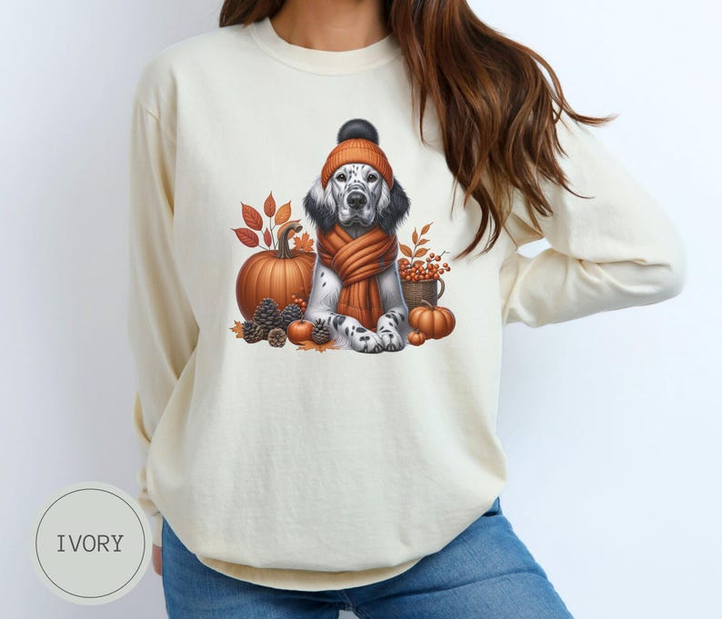 May include: A cream-colored long-sleeved sweatshirt with a graphic of a dog wearing a knitted hat and scarf, surrounded by pumpkins and fall leaves.