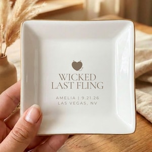 May include: A square white ceramic trinket dish with gold-toned edges. The center features a brown heart icon with small devil horns and the text WICKED LAST FLING, followed by the name AMELIA, the date 9.21.26, and the location LAS VEGAS, NV.