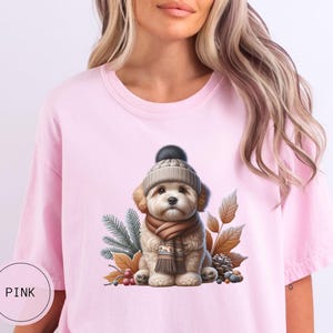 May include: A pink t-shirt with a graphic of a small dog wearing a brown and white knit hat and scarf. The dog is surrounded by autumn leaves and pine cones.