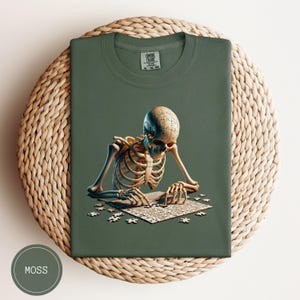 May include: Moss green t-shirt featuring a skeleton solving a jigsaw puzzle. The skeleton's skull is made of puzzle pieces. The shirt has a crew neck and a tag that reads "COMFORT COLORS".