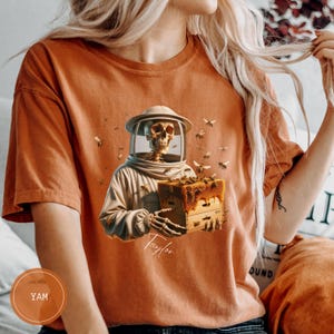 May include: Orange t-shirt with a skeleton beekeeper graphic, holding a honeycomb box, with bees buzzing around. The skeleton wears a beekeeping suit and helmet. The name "Taylor" is written on the shirt.