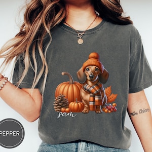 Op de afbeelding: Een grijs T-shirt met een schattig cartoon teckel die een bruine en oranje hoed en sjaal draagt. De teckel is omringd door herfstachtige elementen zoals pompoenen, bladeren en bessen. Het shirt heeft de naam "Sam" erop gedrukt.