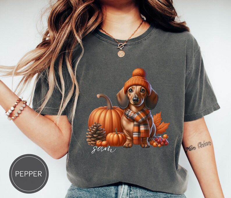 Op de afbeelding: Een grijs T-shirt met een schattig cartoon teckel die een bruine en oranje hoed en sjaal draagt. De teckel is omringd door herfstachtige elementen zoals pompoenen, bladeren en bessen. Het shirt heeft de naam "Sam" erop gedrukt.