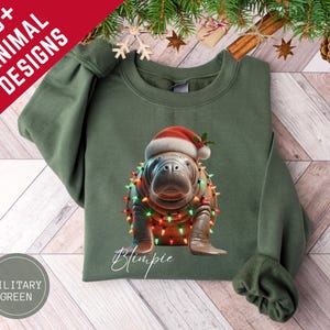 May include: Military green sweatshirt featuring a manatee wearing a Santa hat and Christmas lights. The manatee is named "Blimpie" in a cursive font. The top left corner has the text "20+ ANIMAL DESIGNS" in white.