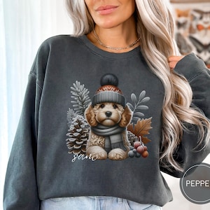 May include: A dark gray sweatshirt with a graphic of a dog wearing a winter hat and scarf. The dog is surrounded by pine cones, berries, and leaves. The text "Sam" is printed below the dog.
