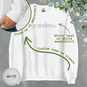 May include: White sweatshirt with the word "grandma" embroidered on the front. The text is in a cursive font and is white. The sweatshirt has a white text label on the sleeve that says "Custom name on sleeve".