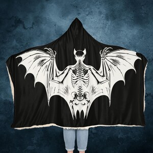 May include: Black hooded blanket with a white skeleton bat design. The bat has large wings and a detailed skeletal structure.