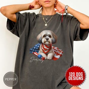 May include: A gray t-shirt with a white and red American flag design featuring a small white dog with a red, white, and blue bandana. The dog's name, "Sammy", is written in cursive below the dog. The t-shirt has a distressed look and is perfect for dog lovers.