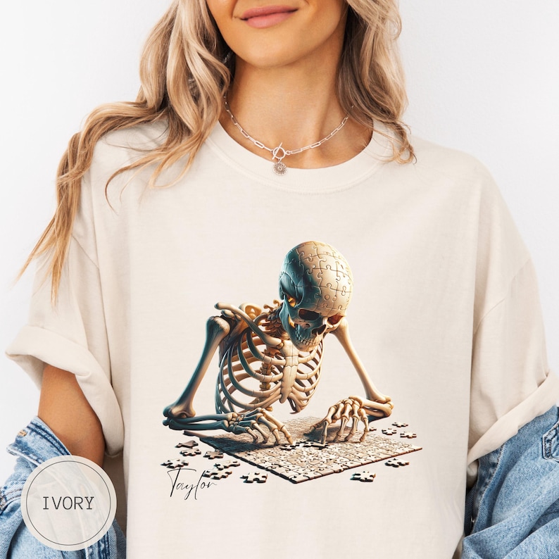 May include: Ivory-colored t-shirt featuring a detailed illustration of a skeleton assembling a jigsaw puzzle. The skeleton's head is a puzzle, and the puzzle pieces are scattered around. The word "Taylor" is written in cursive. The shirt has a relaxed fit.