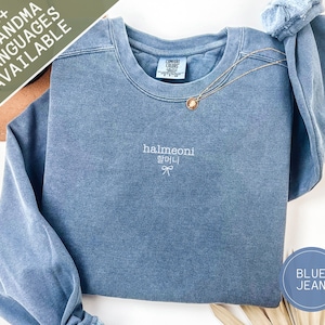 May include: A blue jean-colored sweatshirt with the word "halmeoni" embroidered in white, along with Korean characters and a bow. A gold necklace with a pendant rests on the shirt. The text "30+ Grandma Languages Available" is in the upper left corner.