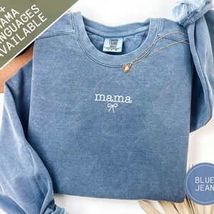 May include: A blue jean-colored sweatshirt with the word "mama" embroidered in white, with a bow underneath. A gold necklace with a pendant rests on the shirt. The text "30+ MAMA LANGUAGES AVAILABLE" is in the upper left corner.