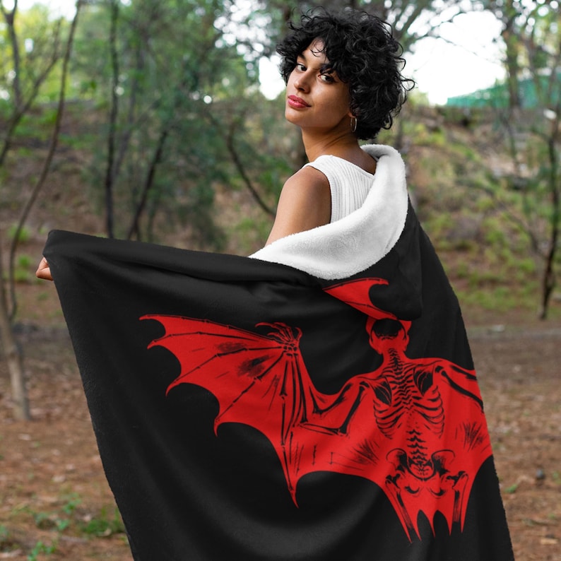 May include: A black hooded blanket with a red skeleton bat design. The blanket is soft and cozy, perfect for cuddling up on the couch.