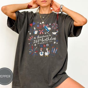 May include: Dark grey t-shirt featuring a graphic celebrating America's 250th birthday. The design includes fireworks, flags, and cartoon geese. The text reads "America's 250th birthday 4th of July 1776-2026".