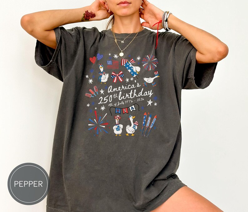 May include: Dark grey t-shirt featuring a graphic celebrating America's 250th birthday. The design includes fireworks, flags, and cartoon geese. The text reads "America's 250th birthday 4th of July 1776-2026".