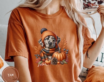 Op de afbeelding: Oranje T-shirt met een afbeelding van een Dalmatiër die een muts en sjaal draagt, omringd door herfstbladeren, pompoenen en bessen. De tekst "Sam" is onder de hond gedrukt.