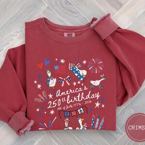 May include: Crimson-coloured sweatshirt with a graphic celebrating America's 250th birthday. The design includes fireworks, flags, and geese wearing patriotic attire. The text reads "America's 250th birthday 4th of July 1776-2026" with "USA" on a banner.