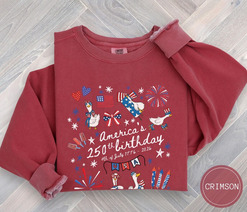 May include: Crimson-coloured sweatshirt with a graphic celebrating America's 250th birthday. The design includes fireworks, flags, and geese wearing patriotic attire. The text reads "America's 250th birthday 4th of July 1776-2026" with "USA" on a banner.