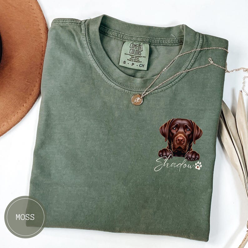 以下が含まれることがあります： モスグリーンのTシャツに、茶色のラブラドールレトリバーが下から顔を出しています。犬の名前「シャドウ」は、犬の下に白い文字で書かれています。Tシャツには「コンフォートカラーズ」と書かれたタグが付いており、小さな銀色のネックレスがTシャツにかけられています。