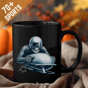 Puede incluir: Taza de cerámica negra con un diseño de esqueleto practicando bobsleigh. El esqueleto lleva casco y traje de carreras. La taza tiene el texto "70+ SPORTS" y la firma "Taylor". El bobsleigh es azul claro.