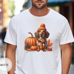 Op de afbeelding: Wit T-shirt met een afbeelding van een teckel die een bruine en oranje gebreide muts en sjaal draagt, omringd door pompoenen en herfstbladeren. De tekst "Fall" is onder de afbeelding gedrukt.