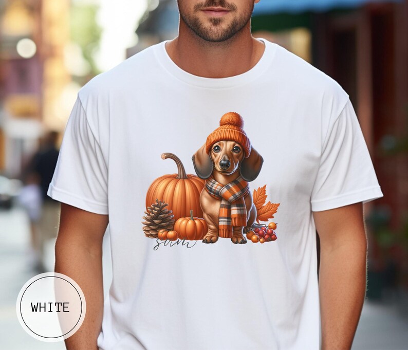 Op de afbeelding: Wit T-shirt met een afbeelding van een teckel die een bruine en oranje gebreide muts en sjaal draagt, omringd door pompoenen en herfstbladeren. De tekst "Fall" is onder de afbeelding gedrukt.