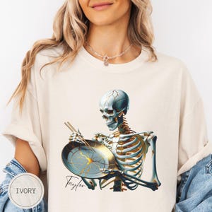 Könnte beinhalten: Elfenbeinfarbenes T-Shirt mit einer detaillierten Illustration eines Skeletts, das eine Bratpfanne mit Essstäbchen hält. Die Pfanne hat ein goldenes Rissmuster. Das Wort "Taylor" ist kursiv geschrieben. Ein lässiges Kleidungsstück für den Alltag.