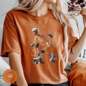 May include: Orange t-shirt with a skeleton roller skating, wearing a white helmet, knee pads, and roller skates. The name "Taylor" is written in cursive. The shirt is a solid orange colour.