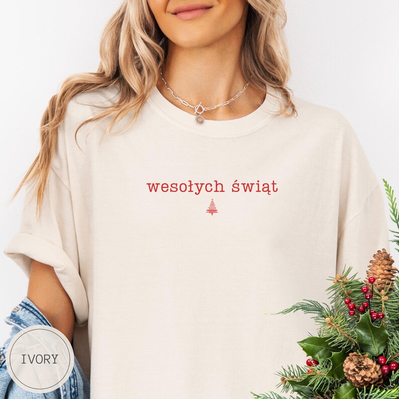 Może przedstawiać: T-shirt z kr&oacute;tkim rękawem w kolorze kości słoniowej z czerwonym napisem "wesołych świąt" wyśrodkowanym na klatce piersiowej, umieszczonym nad małą czerwoną grafiką choinki. Koszulka ma okrągły dekolt i luźny kr&oacute;j, noszona z naszyjnikiem.
