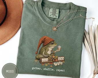 May include: A moss green t-shirt featuring a graphic of a frog wearing an orange knit cap, holding a coffee cup, and sitting beside mahjong tiles. The text "gather, shuffle, repeat" is printed below the graphic. The shirt has a Comfort Colors label.