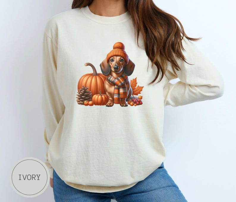 Op de afbeelding: Een wit longsleeve sweatshirt met een schattige illustratie van een teckel die een bruine en oranje hoed en sjaal draagt, omringd door pompoenen en herfstbladeren.