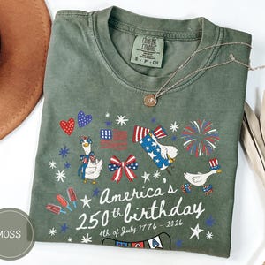May include: Moss green t-shirt with a patriotic design celebrating America's 250th birthday. The graphic includes cartoon geese, fireworks, and the American flag. The text reads "America's 250th birthday 4th of July 1776-2026."
