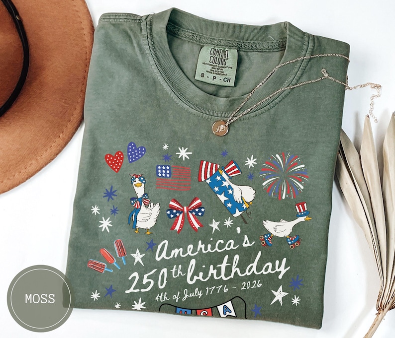 May include: Moss green t-shirt with a patriotic design celebrating America's 250th birthday. The graphic includes cartoon geese, fireworks, and the American flag. The text reads "America's 250th birthday 4th of July 1776-2026."