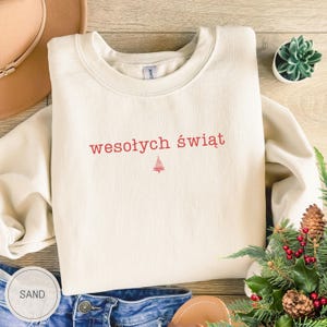 Może przedstawiać: Bluza z okrągłym dekoltem w kolorze piaskowym z wyhaftowanym na czerwono napisem "wesołych świąt" na środku klatki piersiowej, umieszczonym nad małą, minimalistyczną czerwoną ikoną choinki. Odzież posiada długie rękawy i prążkowany dekolt.