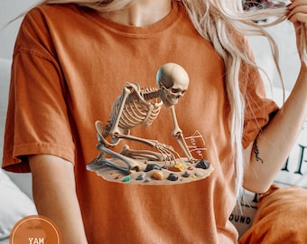 May include: Burnt orange t-shirt featuring a detailed skeleton graphic kneeling on sand, surrounded by colorful stones. The skeleton appears to be searching or working. The shirt has a relaxed fit and short sleeves. The text "Taylor" is visible on the graphic.