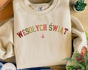 Może przedstawiać: Bluza z okrągłym dekoltem w kolorze piaskowym z napisem WESOŁYCH ŚWIĄT w łukowatej, wielokolorowej czcionce, z małą czerwoną ikoną choinki wyśrodkowaną pod tekstem. Odzież posiada długie rękawy oraz ściągacze przy dekolcie, mankietach i u dołu.