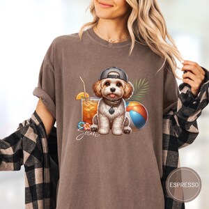 May include: A brown t-shirt with a cartoon image of a dog wearing a baseball cap and holding a beach ball. The dog is sitting next to a glass of orange juice and a tropical flower. The text "Sam" is printed below the image.