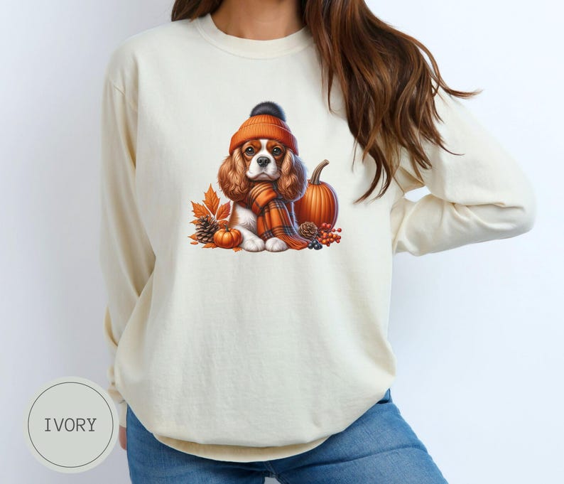 May include: A cream-colored long-sleeved sweatshirt with a graphic of a Cavalier King Charles Spaniel wearing a brown and orange hat and scarf, surrounded by fall leaves and a pumpkin.