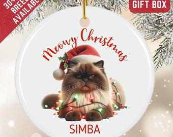 May include: A round white ceramic ornament featuring a fluffy brown and cream cat wearing a red Santa hat with holly. The cat is tangled in colorful Christmas lights. The text reads Meowy Christmas and SIMBA in red lettering with a gold string.