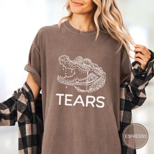 May include: An espresso-colored t-shirt featuring a white line-art graphic of an alligator head with an open mouth and the word TEARS printed in white capital letters below the illustration. The shirt has a crew neckline and short sleeves.