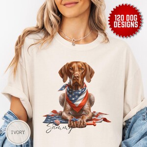 May include: A white t-shirt featuring a brown and white dog wearing a red, white and blue bandana. The dog is sitting on a red, white and blue American flag. The dog's name, "Sammy", is written in cursive below the dog. The shirt is labelled "IVORY".