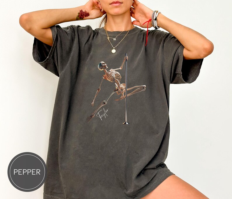 May include: Dark grey t-shirt featuring a skeleton pole dancing. The skeleton is holding onto a silver pole. The word "Taylor" is written in cursive below the skeleton. The shirt has a relaxed fit.