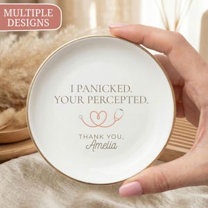 May include: A round white ceramic trinket dish with a gold-coloured rim. The centre features the text I PANICKED. YOUR PERCEPTED. above a pink line-art stethoscope shaped like a heart, followed by the text THANK YOU, Amelia in script font.