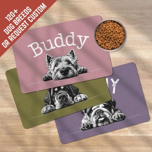 May include: Three dog-themed placemats in pink, green, and purple, each with a different dog breed illustration. The pink mat has the name "Buddy" in white. A metal bowl filled with dog food sits at the top. Text reads "120+ DOG BREEDS OR REQUEST CUSTOM".