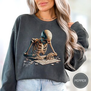 May include: Dark gray sweatshirt featuring a detailed illustration of a skeleton assembling a jigsaw puzzle. The skeleton's skull is also a puzzle. The name "Taylor" is printed in cursive. The sweatshirt has a Pepper label.