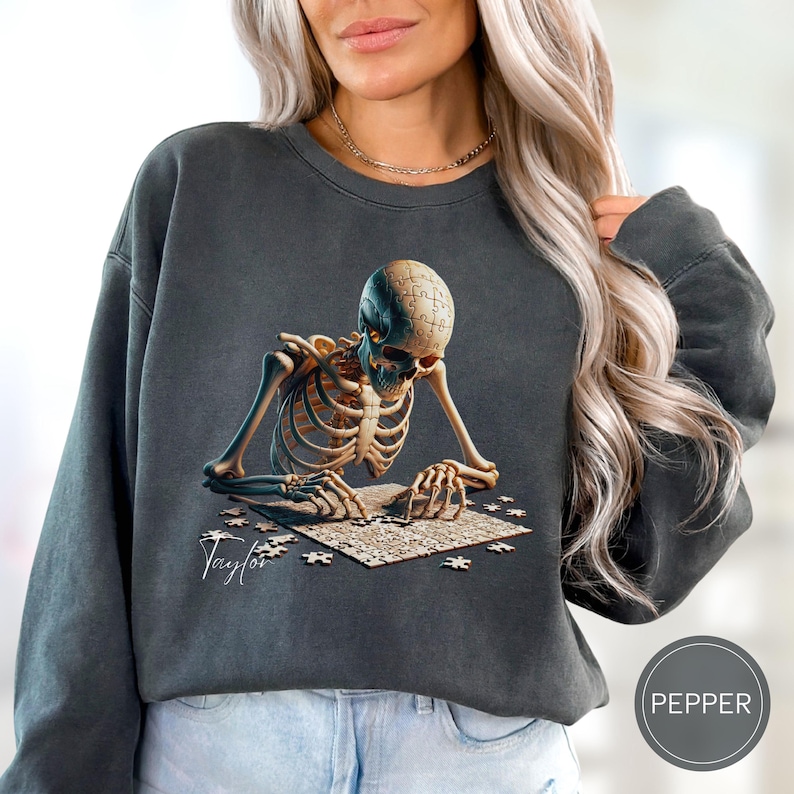May include: Dark gray sweatshirt featuring a detailed illustration of a skeleton assembling a jigsaw puzzle. The skeleton's skull is also a puzzle. The name "Taylor" is printed in cursive. The sweatshirt has a Pepper label.